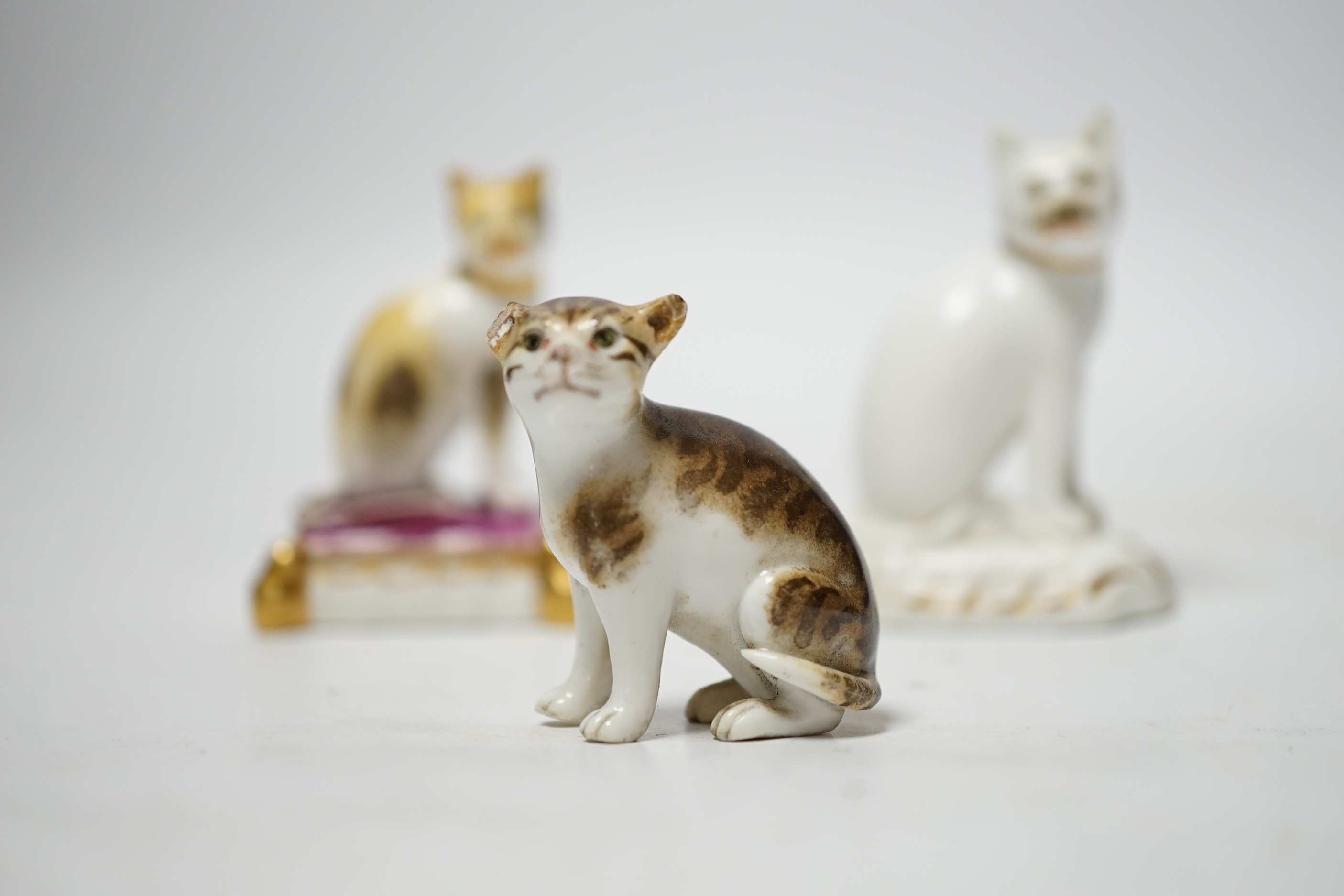 Two Rockingham porcelain models of seated cats, c.1830, the cat seated on a maroon cushion with impressed Rockingham Mark and incised model number 17, 5.3cm high, the white and gilt example with incised model number 11,